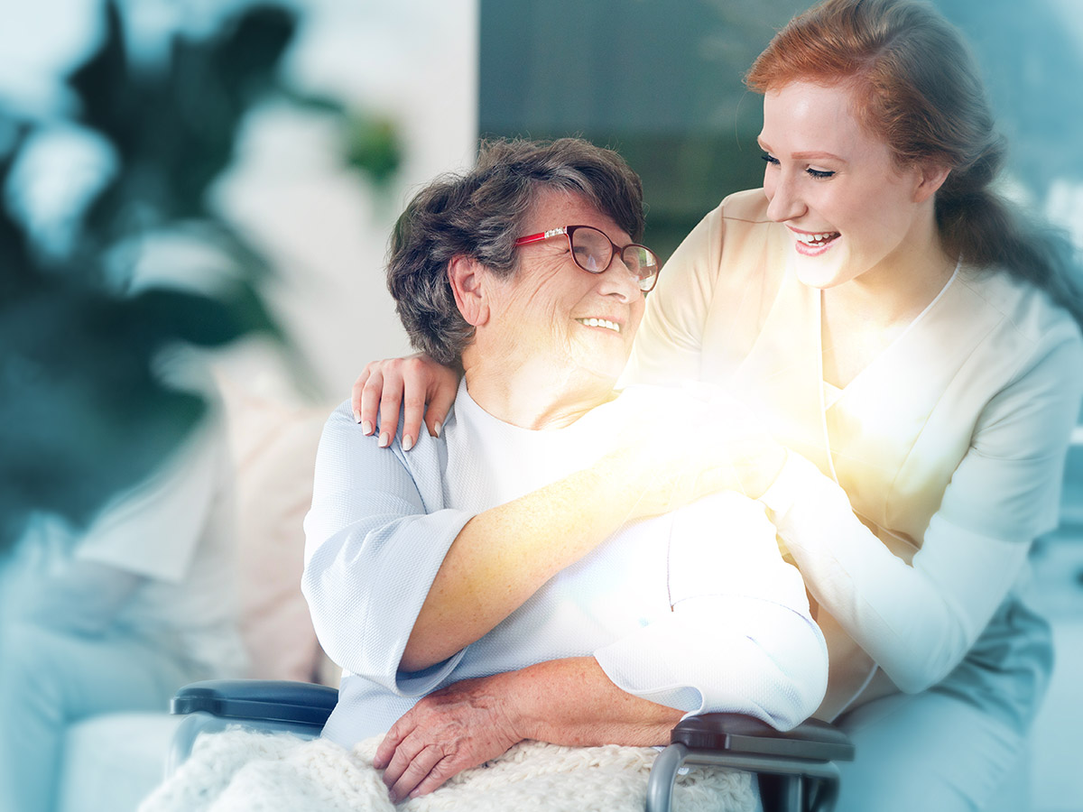 Older female patient in wheelchair smiling to her caregiver who is smiling back and holding her hand